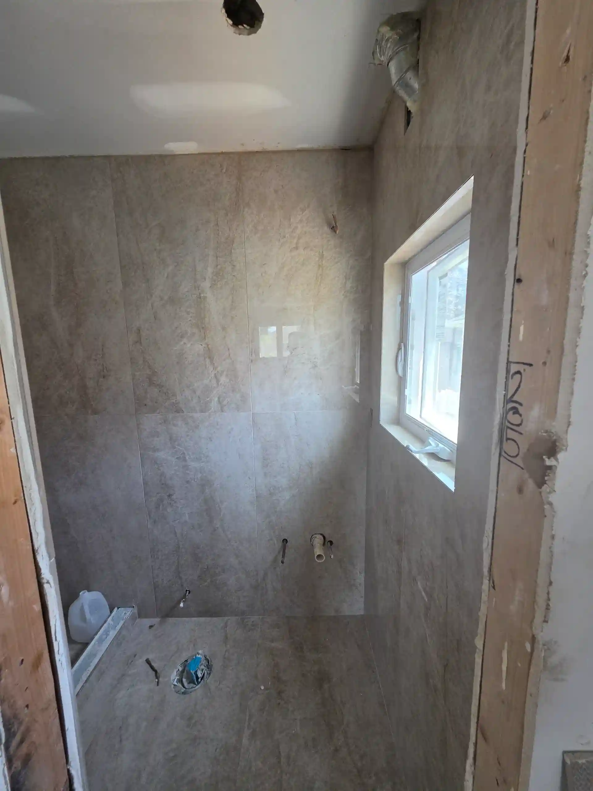 Bathroom renovation in progress with gray marble-look tile, window and exposed plumbing in Brooklyn NY